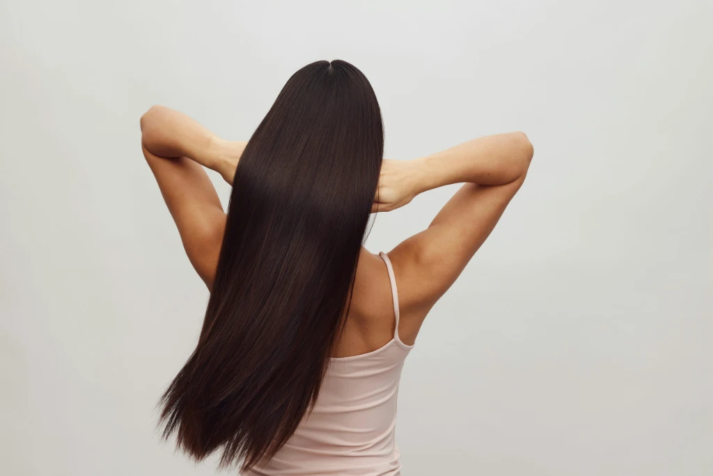 Back view of a woman with smooth, shiny, long brunette hair demonstrating healthy hair growth and strength against a neutral background.