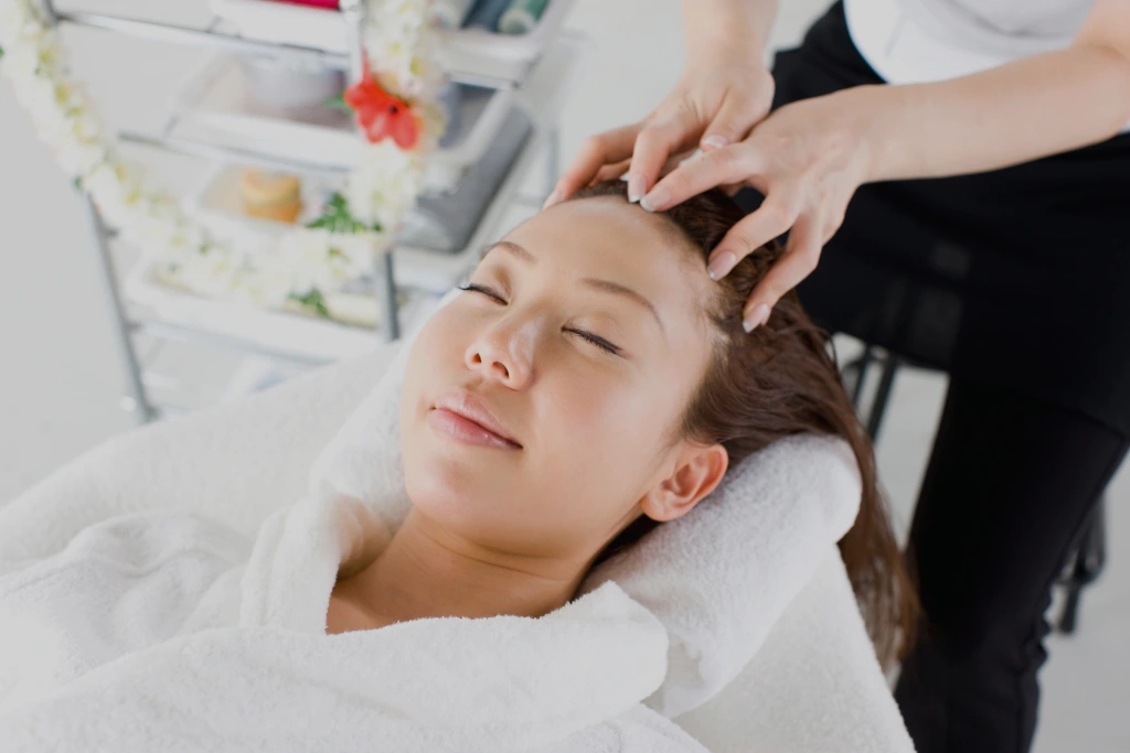 A close-up, high-angle view of a woman with her eyes closed receiving a professional head and scalp massage in a bright, clean wellness spa setting.