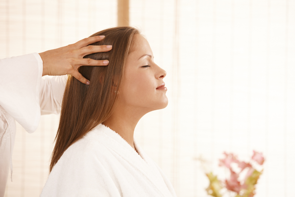 A woman in a white robe with her eyes closed in a profile view, receiving a professional scalp massage in a bright, tranquil spa setting.
