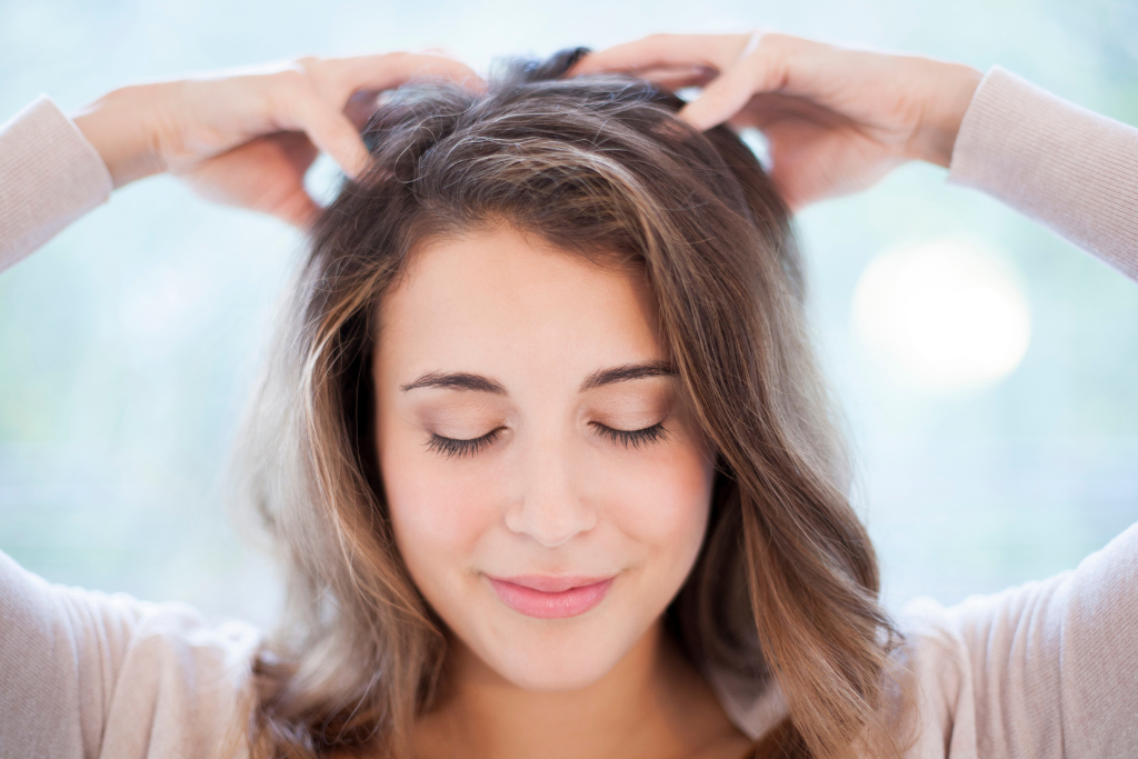 A woman with her eyes closed smiling peacefully while massaging her head with both hands, set against a soft, blurred outdoor background.