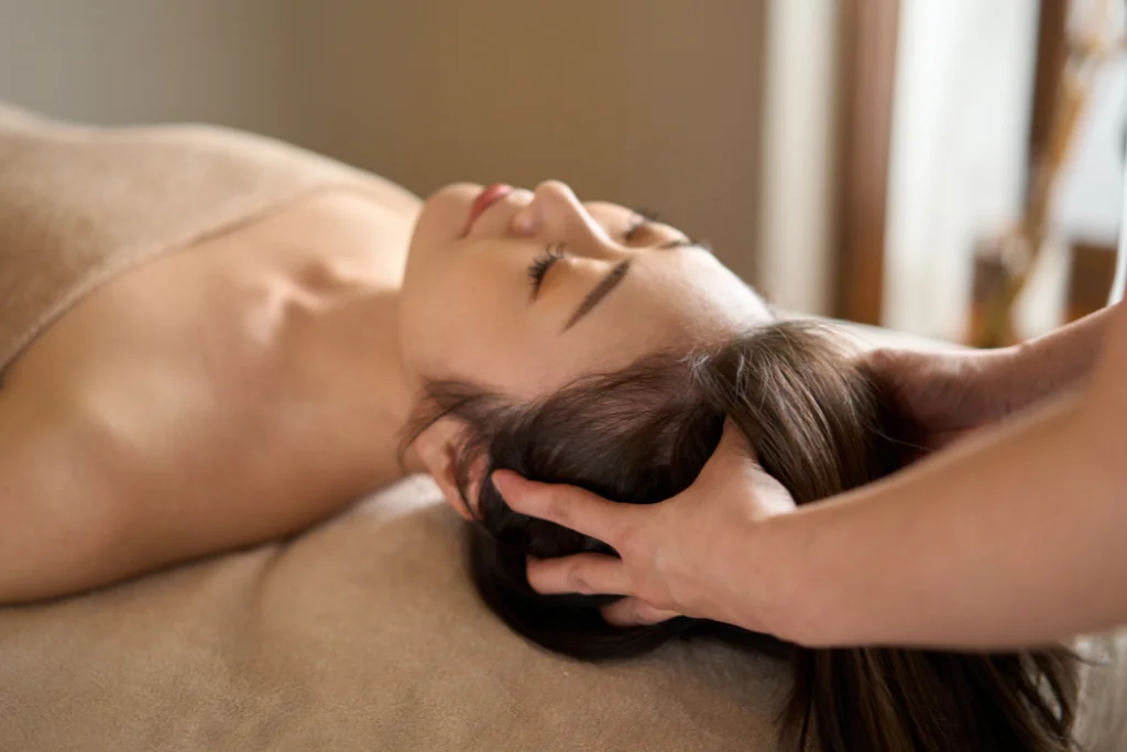 A woman lying on a massage table with her eyes closed, receiving a relaxing head and scalp massage from a professional therapist in a spa setting.