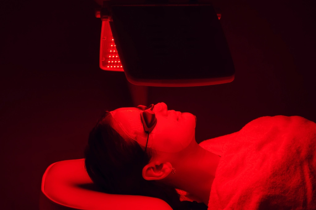A person wearing protective eye goggles receiving a red light therapy facial treatment in a dark room; photorealistic close-up of a professional LED light panel emitting red light over a client's face.