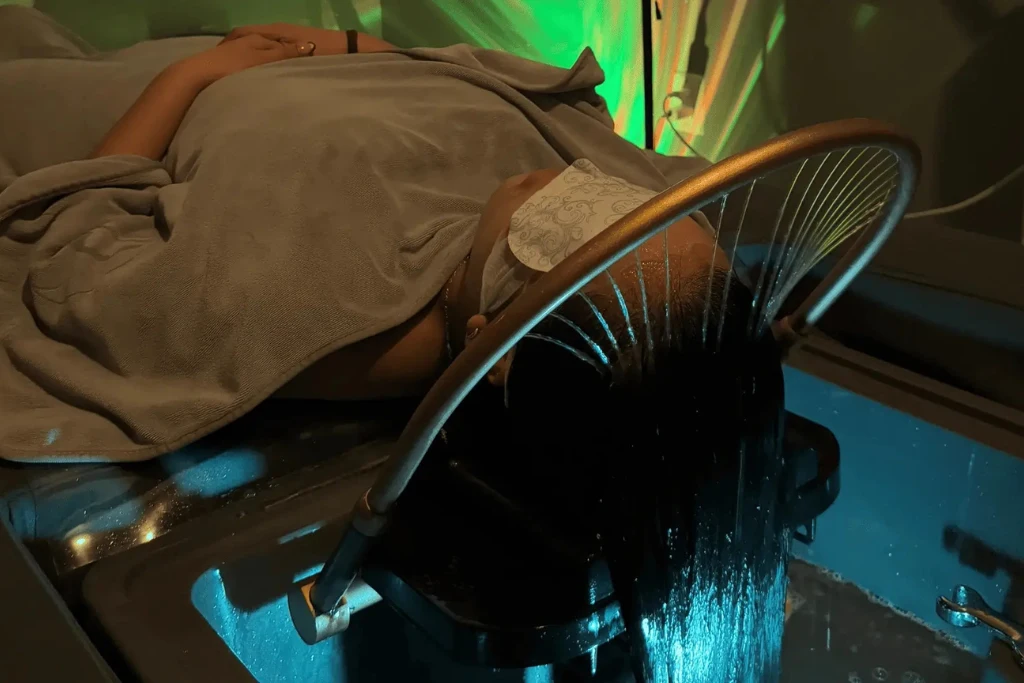 A person lies face-up on a spa bed, covered in a grey towel with an eye mask on, while a circular "water halo" device sprays gentle streams of water over their hair and scalp into a basin.