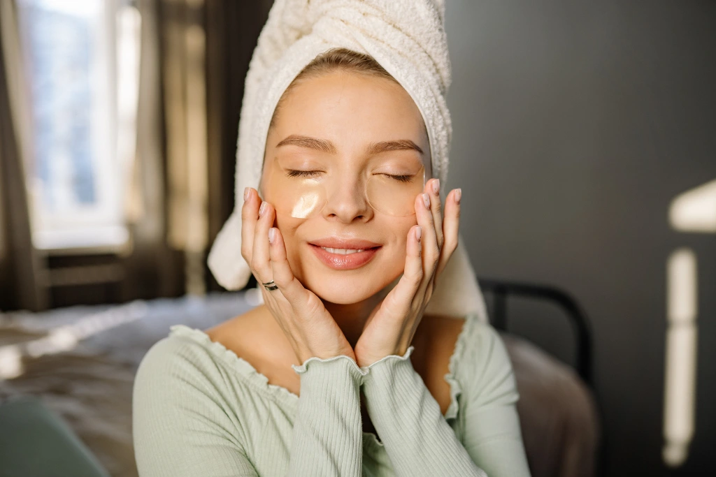 A woman with a towel-wrapped head smiling while performing a gentle facial massage at home as she applies gold hydrogel eye patches.