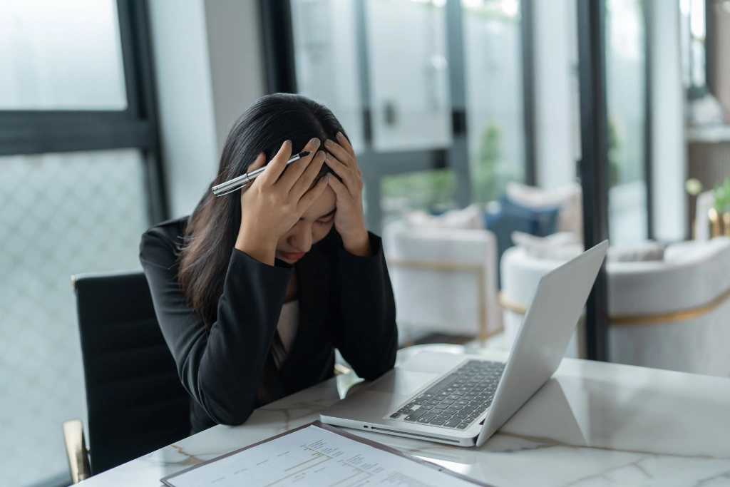A distressed businesswoman in a black blazer at her desk, holding her head in frustration, with a laptop, documents, and an office background.