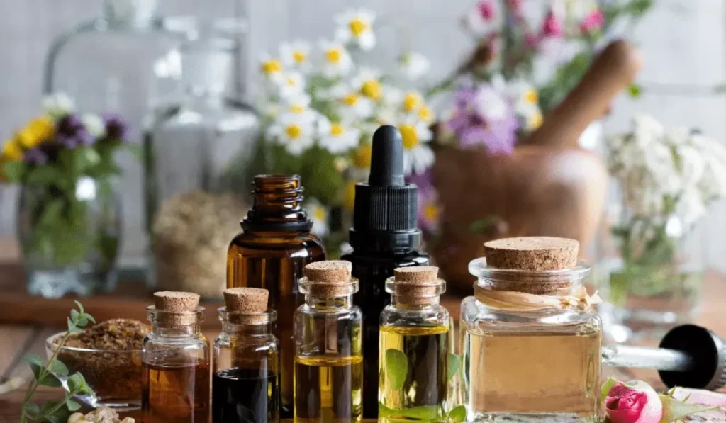 A still life image featuring a collection of small glass bottles filled with various colored essential oils, arranged on a wooden surface. The bottles have cork stoppers and are of different sizes and shapes. In the background, there are more glass bottles, dried herbs, flowers in vases, and a wooden mortar and pestle, suggesting a natural or apothecary setting. The lighting is soft and natural, highlighting the textures of the wood and glass.